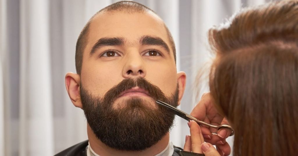 Professional barber with scissors trimming beard